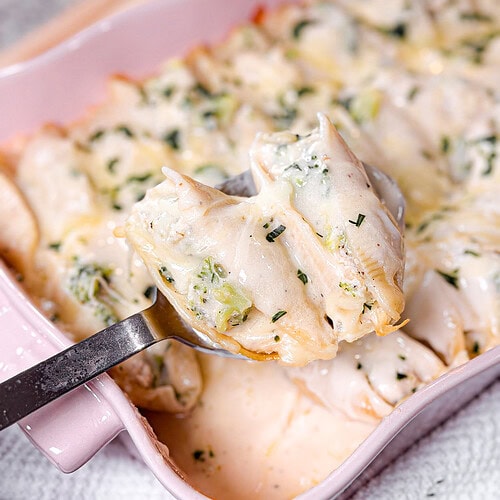 A close-up of Chicken Alfredo Stuffed Shells filled with a creamy mixture, topped with sauce and herbs, in a pink baking dish. A spoon lifts several shells out of the dish.