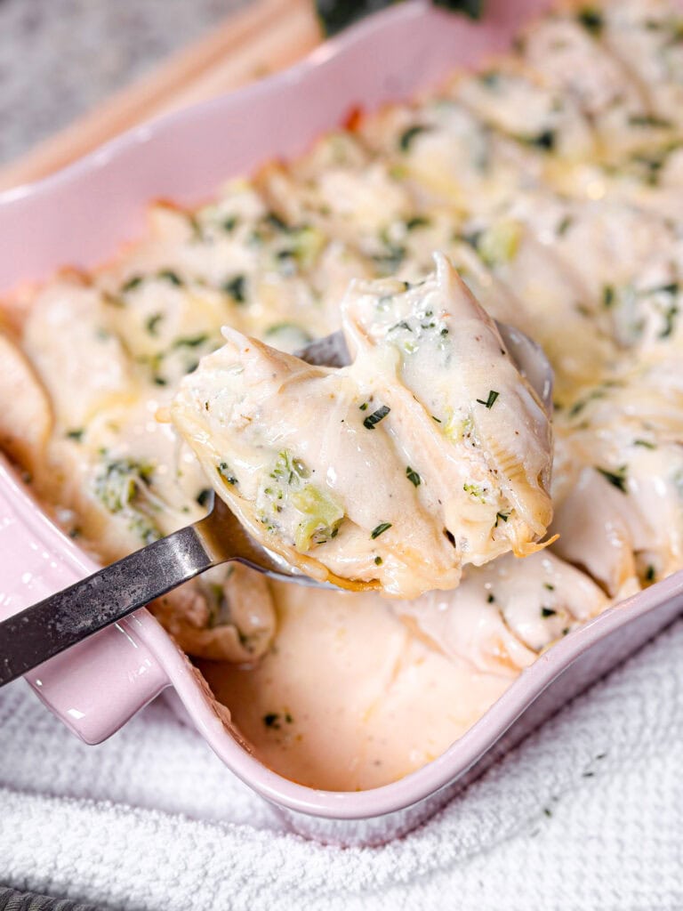 A close-up of Chicken Alfredo Stuffed Shells filled with a creamy mixture, topped with sauce and herbs, in a pink baking dish. A spoon lifts several shells out of the dish.
