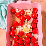 Pink baking dish with whole cherry tomatoes, sliced onions, garlic, and a round piece of seasoned cheese in the center—perfect for making Baked Boursin Cheese Pasta. A light blue towel is nearby on a wooden surface. Ingredients are uncooked and ready to roast.