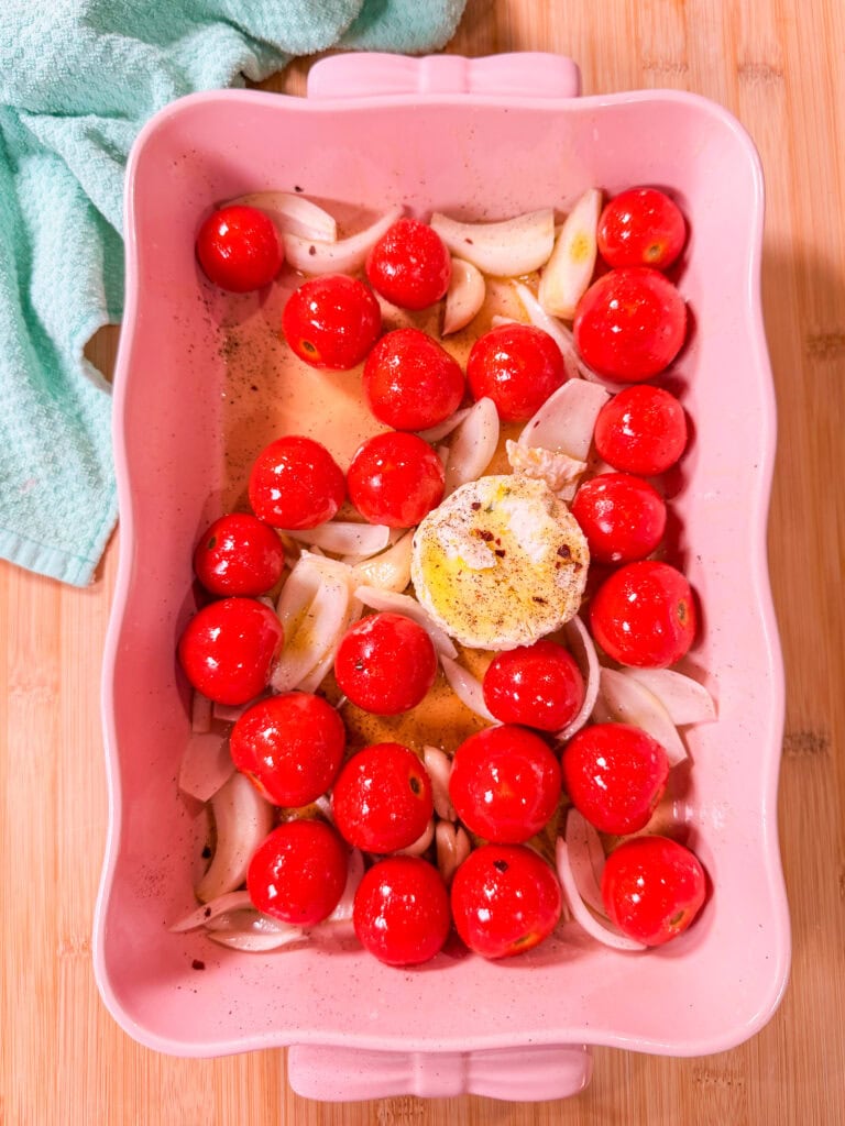 Pink baking dish with whole cherry tomatoes, sliced onions, garlic, and a round piece of seasoned cheese in the center—perfect for making Baked Boursin Cheese Pasta. A light blue towel is nearby on a wooden surface. Ingredients are uncooked and ready to roast.