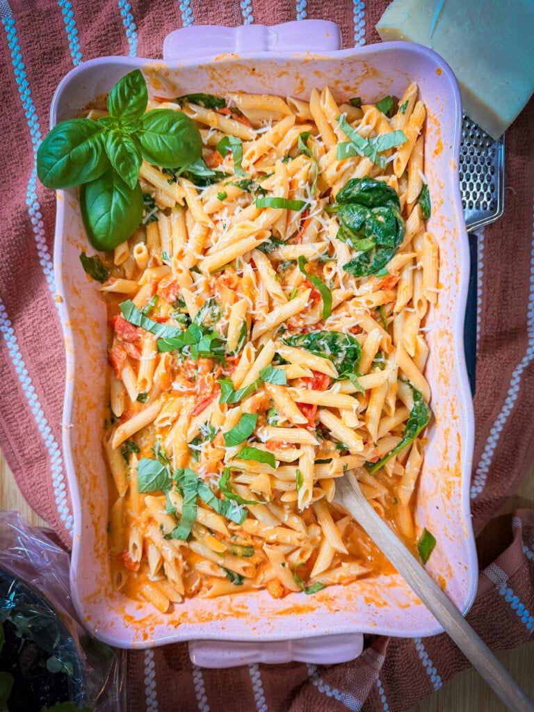 Baked Boursin Cheese Pasta in a pink casserole dish with creamy tomato sauce, spinach, grated cheese, and fresh basil, placed on a striped towel with a wooden spoon and grated cheese nearby.