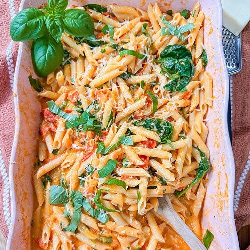 Baked Boursin Cheese Pasta with penne in a creamy tomato sauce and spinach, topped with fresh basil and grated cheese, served in a pink casserole dish with a wooden spoon.