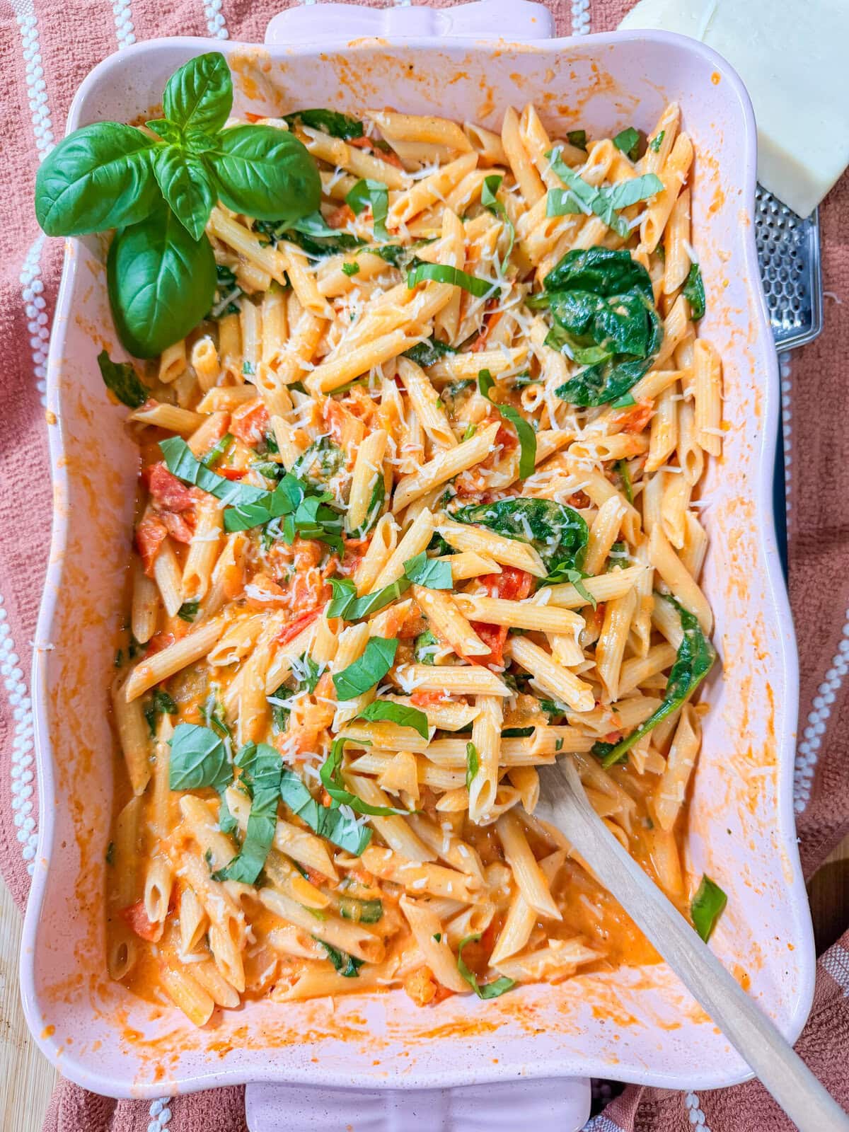 Baked Boursin Cheese Pasta with penne in a creamy tomato sauce and spinach, topped with fresh basil and grated cheese, served in a pink casserole dish with a wooden spoon.