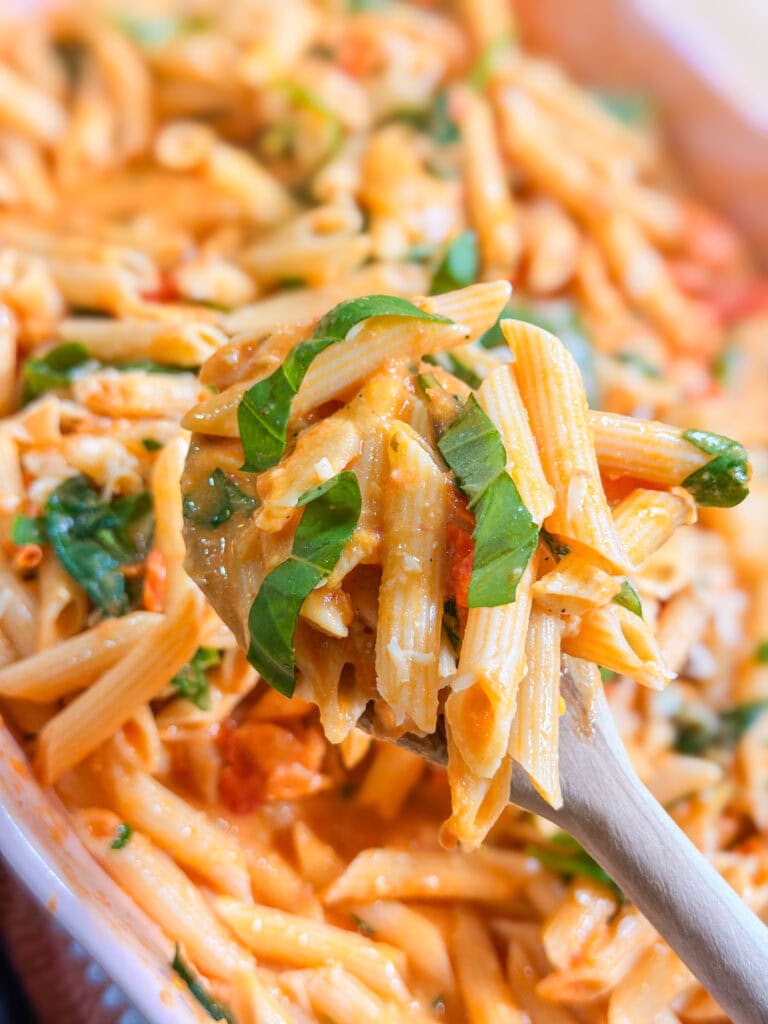 A close-up of creamy Baked Boursin Cheese Pasta with tangy tomato sauce, fresh basil, and melted cheese on a wooden spoon, with more pasta in the background.