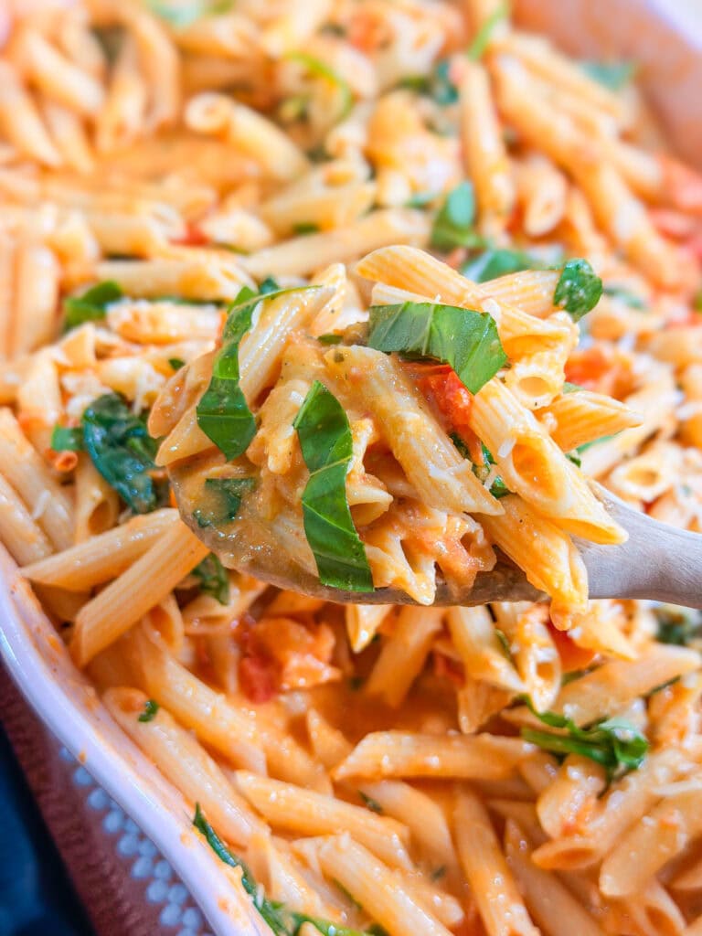 A wooden spoon holds creamy penne pasta with fresh basil, spinach, and cherry tomatoes above a dish filled with the same Baked Boursin Cheese Pasta.