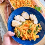 A hand holds a blue plate with sliced chicken breast and creamy tomato penne pasta with spinach, reminiscent of Baked Boursin Cheese Pasta; a casserole dish of pasta and a towel are in the background.