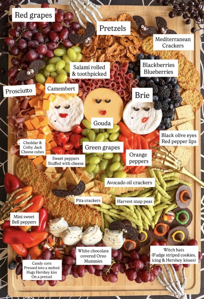 A Halloween-themed charcuterie board with cheeses, meats, crackers, fruits, veggies, and festive treats, neatly labeled. Starring three cheese faces, pretzels, grapes, olives, and sweet and savory snacks on a wooden tray—perfect for anyone searching for easy Halloween food ideas.