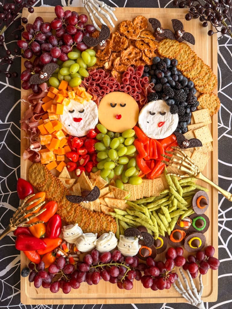 A Halloween-themed charcuterie board, perfect for anyone seeking Easy Halloween Food Ideas, features cheeses shaped as three women’s faces, surrounded by fruit, crackers, pretzels, peppers, and festive Halloween candies—all artfully arranged on a wooden tray decorated with playful spiderwebs.