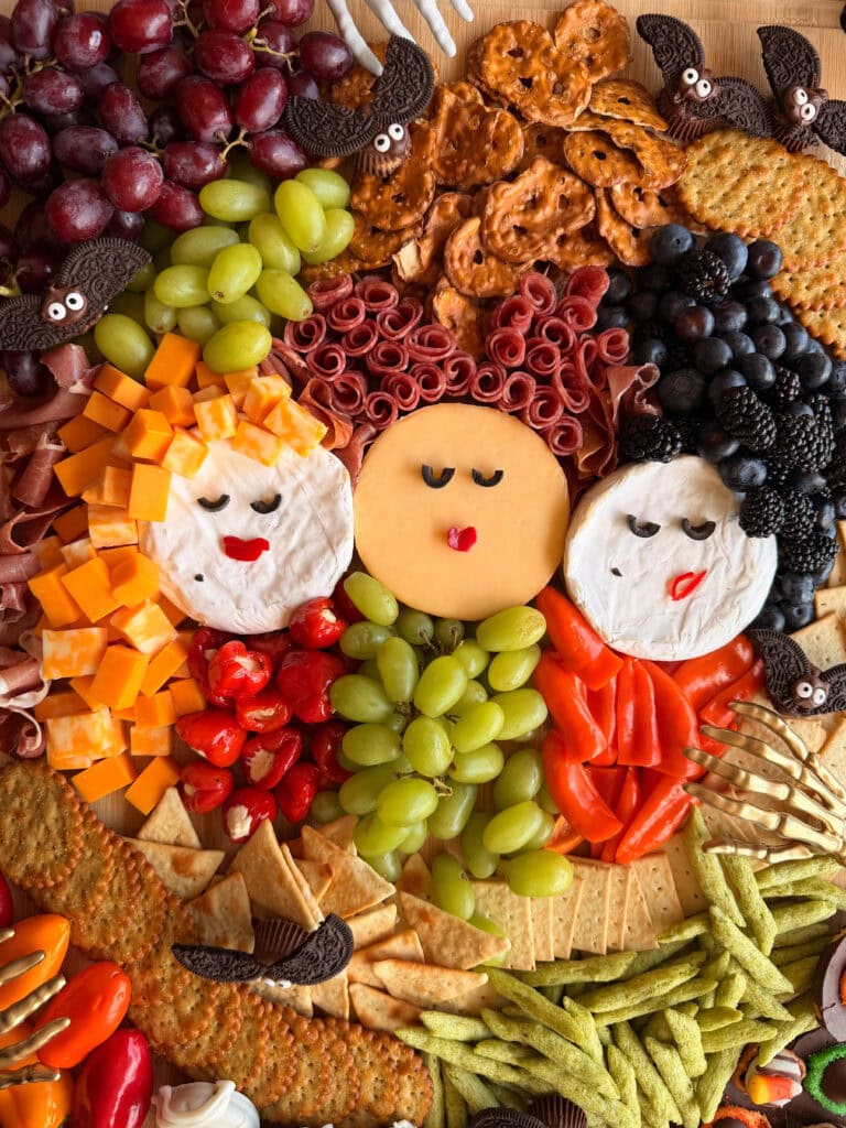 A festive charcuterie board arranged with meats, cheeses, fruits, and crackers to resemble three women’s faces surrounded by colorful food, complete with bats made from cookies and googly eyes—this display is a standout among easy Halloween food ideas.