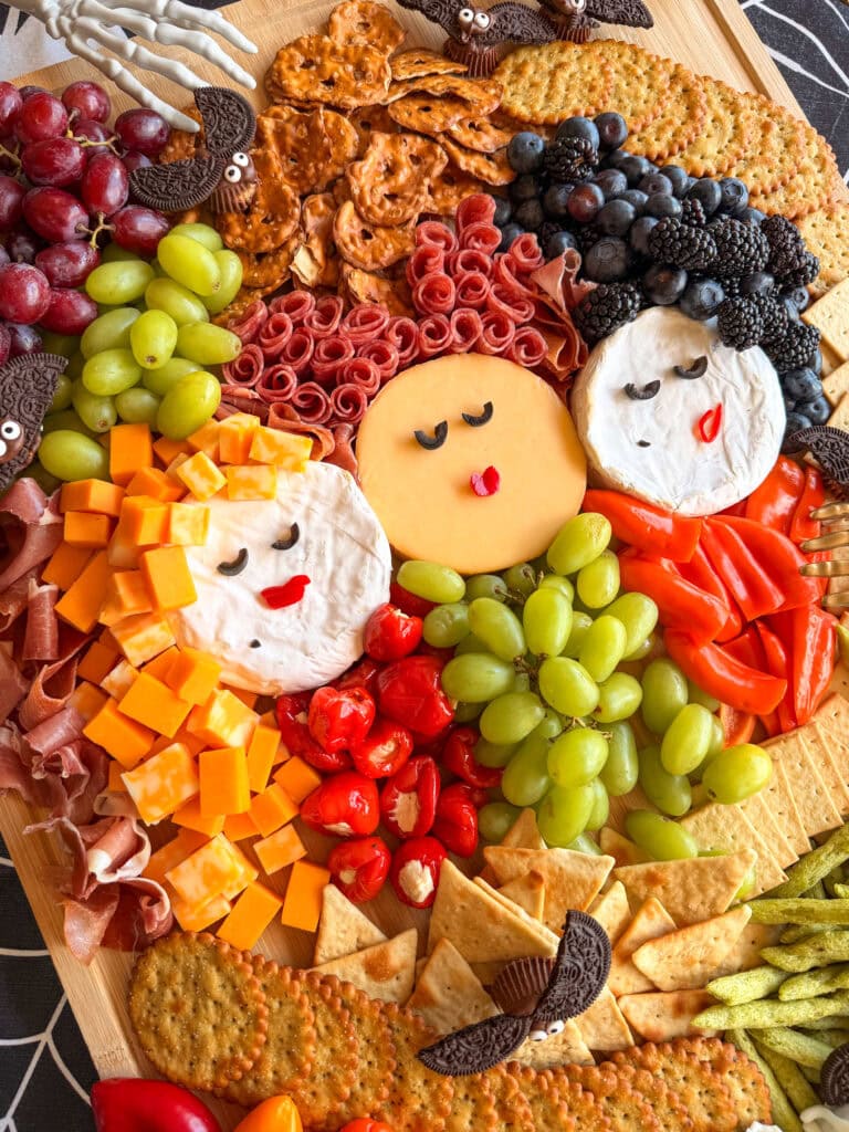 Festive charcuterie board arranged to resemble three women’s faces with cheese, meat, fruit, and crackers—an easy Halloween food idea—featuring whimsical details like dyed cheese hair and cookies shaped like bats.