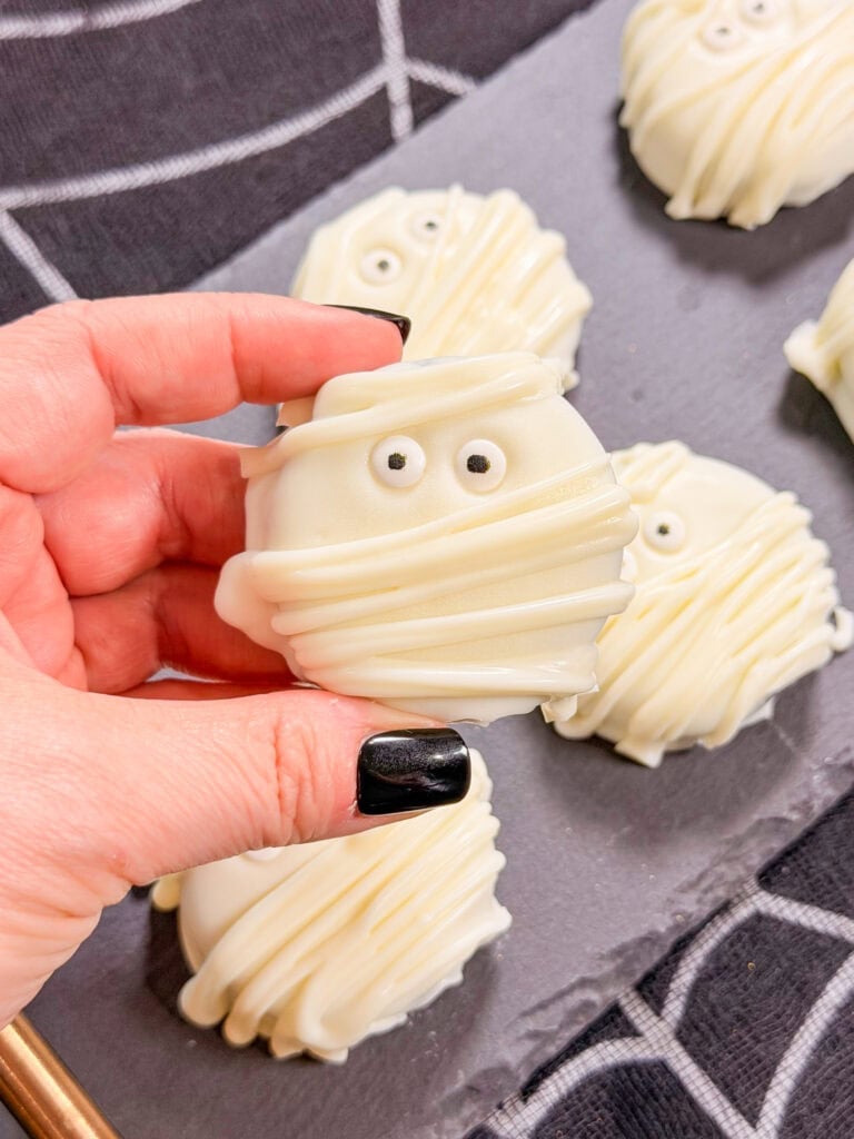 A hand with black nail polish holds a cookie decorated like a mummy with white icing and candy eyes; more mummy cookies—one of the best easy Halloween food ideas—are arranged on a slate tray in the background.
