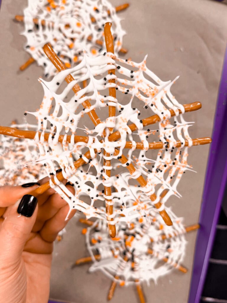 A hand with black nail polish holds a spiderweb-shaped treat made from pretzel sticks and white chocolate, decorated with sprinkles—one of the easy Halloween food ideas perfect for spooky gatherings. More treats are visible on a baking tray in the background.