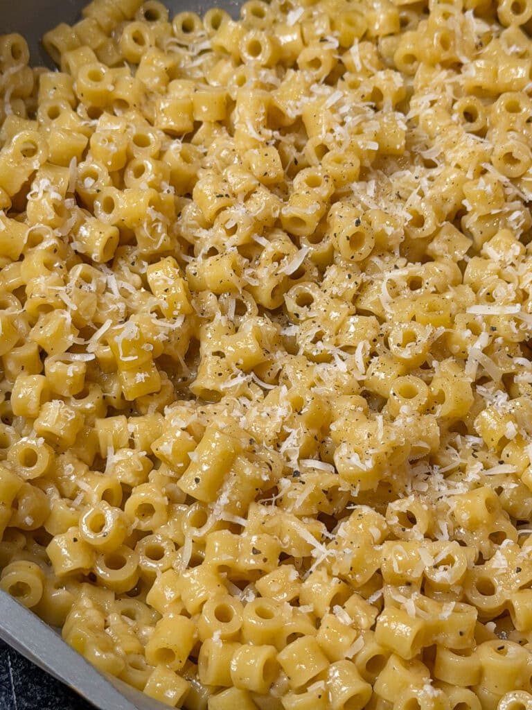 A close-up of cooked ditalini pasta coated in a creamy yellow sauce, just like The Best Buttered Noodles, topped with freshly grated cheese and cracked black pepper.