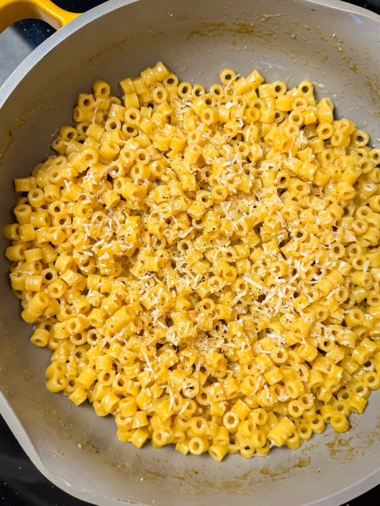 A large skillet filled with small tube-shaped pasta coated in a creamy yellow cheese sauce, reminiscent of The Best Buttered Noodles, topped with freshly grated cheese and cracked black pepper.