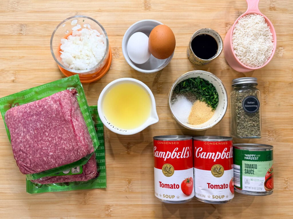 Porcupine Meatballs ingredients: ground beef, chopped onions, two eggs, soy sauce, uncooked white rice, tomato soup cans, tomato sauce can, broth, parsley, garlic powder, salt, and Italian seasoning on a wooden surface.