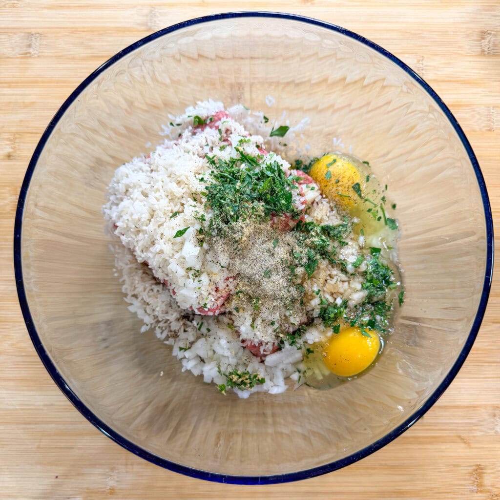 A glass bowl containing raw ground meat, rice, chopped onion, two eggs, and green herbs sits on a wooden surface—ingredients perfectly prepped for classic Porcupine Meatballs.