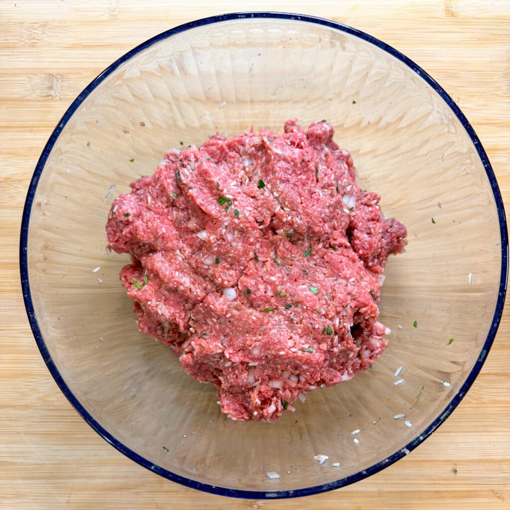 A clear glass bowl filled with seasoned raw ground meat for Porcupine Meatballs, mixed with visible herbs and small pieces of onion, sits on a light wooden surface.
