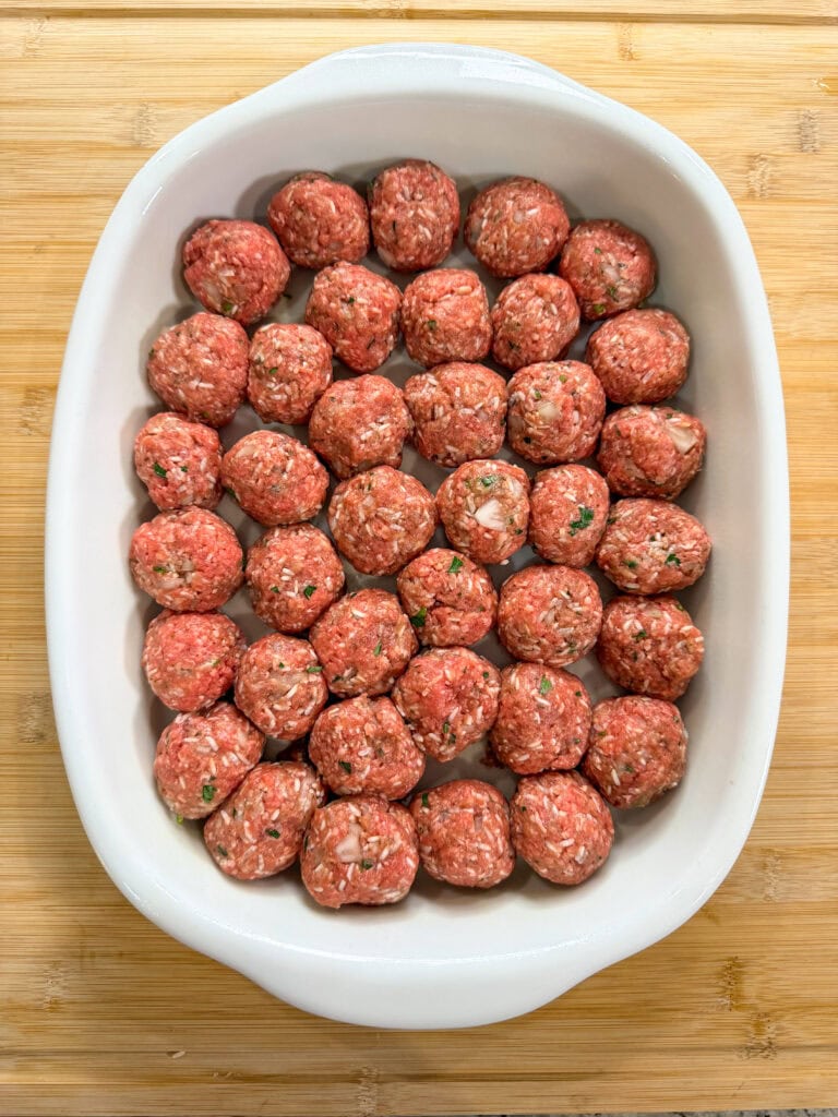 A white baking dish filled with rows of uncooked Porcupine Meatballs—made from ground meat, herbs, diced onions, and rice—rests on a wooden surface.