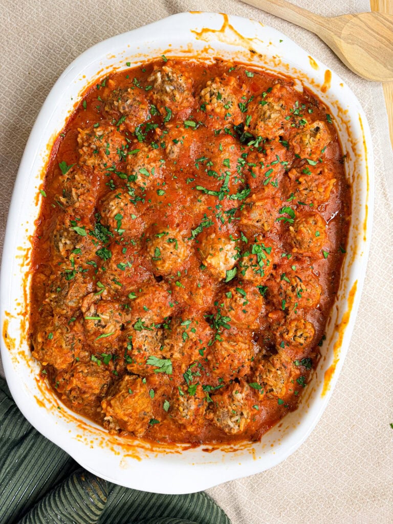 A white baking dish filled with Porcupine Meatballs covered in tomato sauce, topped with chopped fresh herbs, sits on a table with wooden utensils beside it.