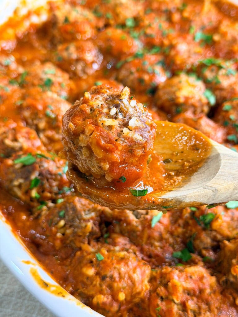 A close-up of a wooden spoon holding a Porcupine Meatball covered in tomato sauce, with a dish full of saucy meatballs and sprinkled herbs in the background.