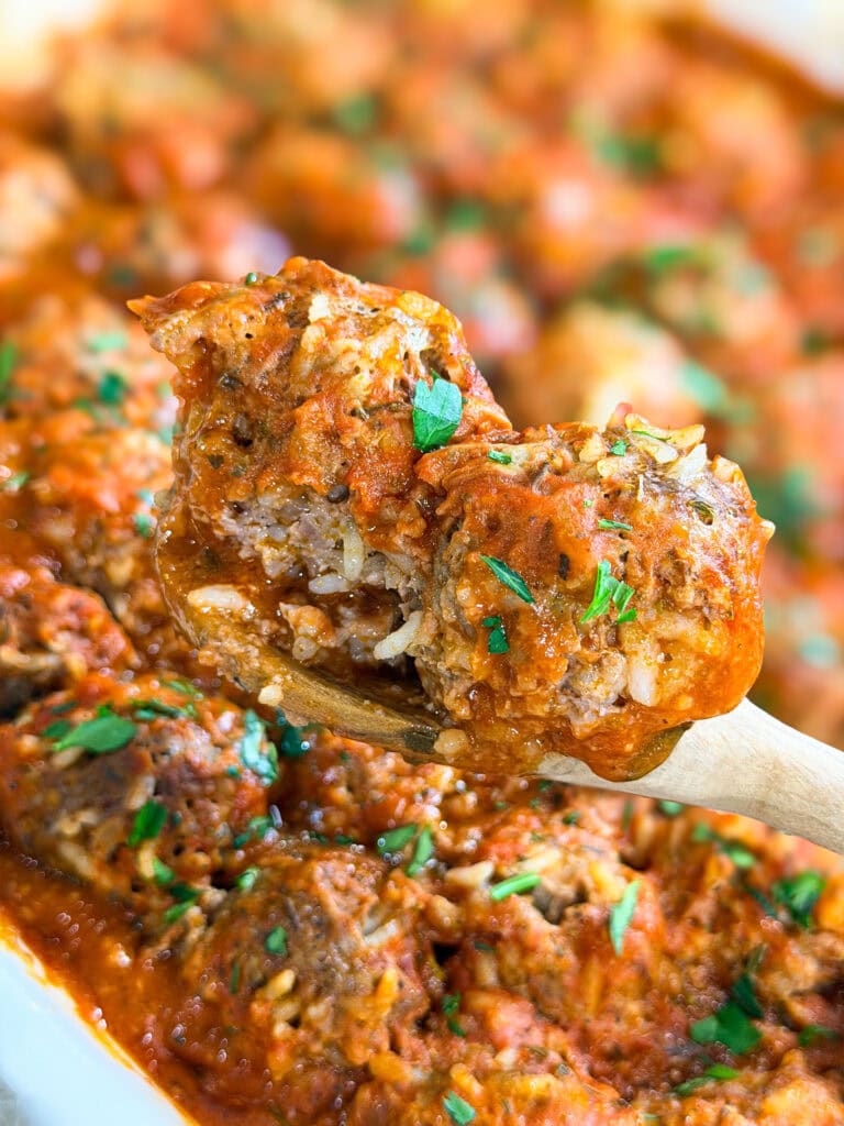 A close-up of a wooden spoon holding Porcupine Meatballs covered in tomato sauce and garnished with chopped parsley, with more meatballs and sauce in the background.