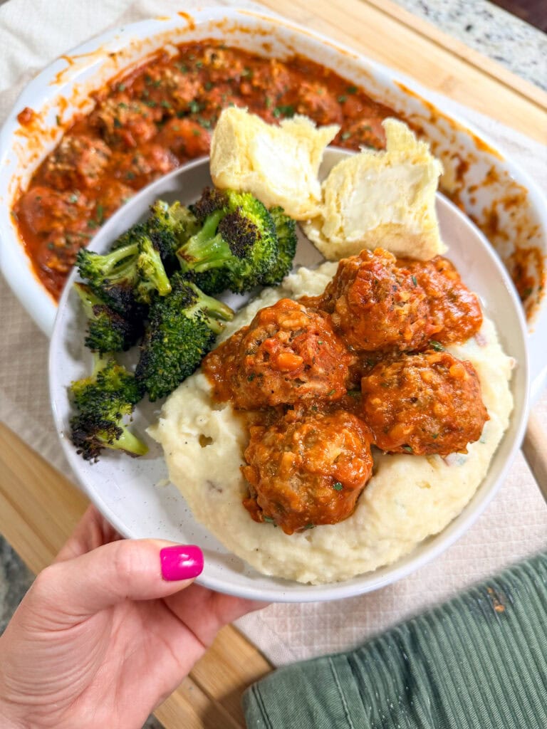 A hand holds a plate with mashed potatoes topped with Porcupine Meatballs in tomato sauce, roasted broccoli, and a buttered bread roll. In the background, a baking dish filled with more meatballs simmers in rich sauce.