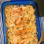 A blue glass baking dish filled with Creamy Chicken and Rice Casserole features baked chicken breasts atop creamy rice with mushrooms, all sprinkled with seasoning. A wooden spoon rests nearby on a striped cloth.