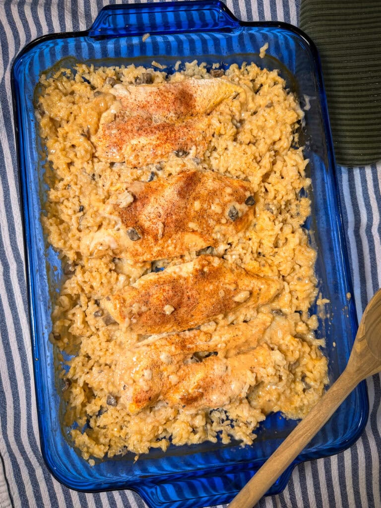 A blue glass baking dish filled with Creamy Chicken and Rice Casserole features baked chicken breasts atop creamy rice with mushrooms, all sprinkled with seasoning. A wooden spoon rests nearby on a striped cloth.