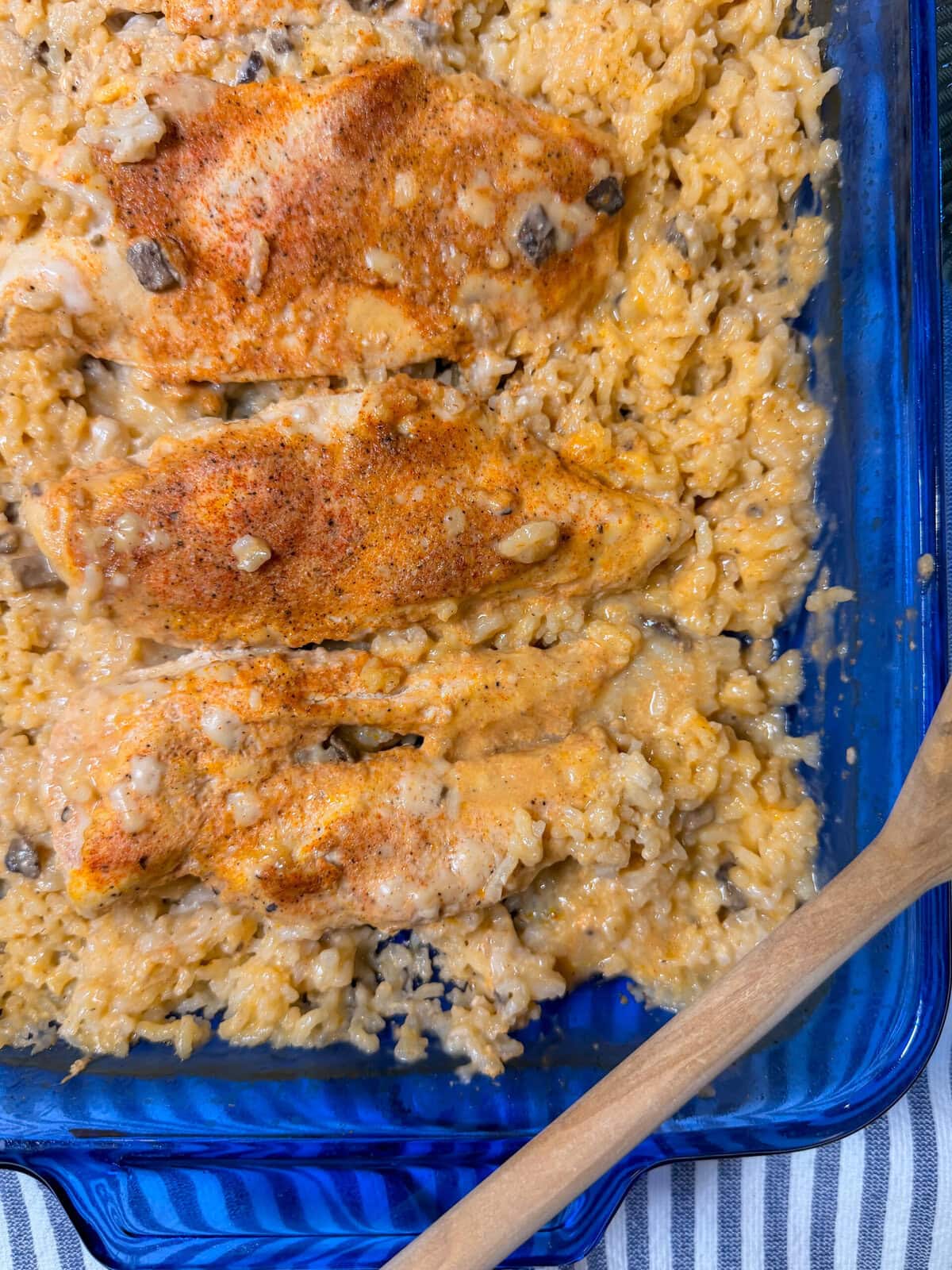 Creamy Chicken and Rice Casserole features baked chicken breasts seasoned with spices, nestled on a bed of creamy rice and mushrooms in a blue glass baking dish, with a wooden spoon on the side.