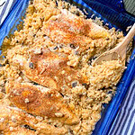 A blue baking dish filled with Creamy Chicken and Rice Casserole, topped with a sprinkle of seasoning. A wooden spoon is scooping up some of the creamy rice, and a striped cloth is partially visible underneath the dish.