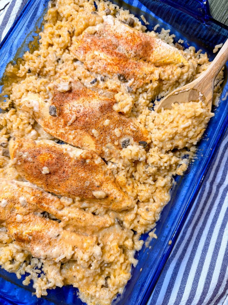 A blue baking dish filled with Creamy Chicken and Rice Casserole, topped with a sprinkle of seasoning. A wooden spoon is scooping up some of the creamy rice, and a striped cloth is partially visible underneath the dish.