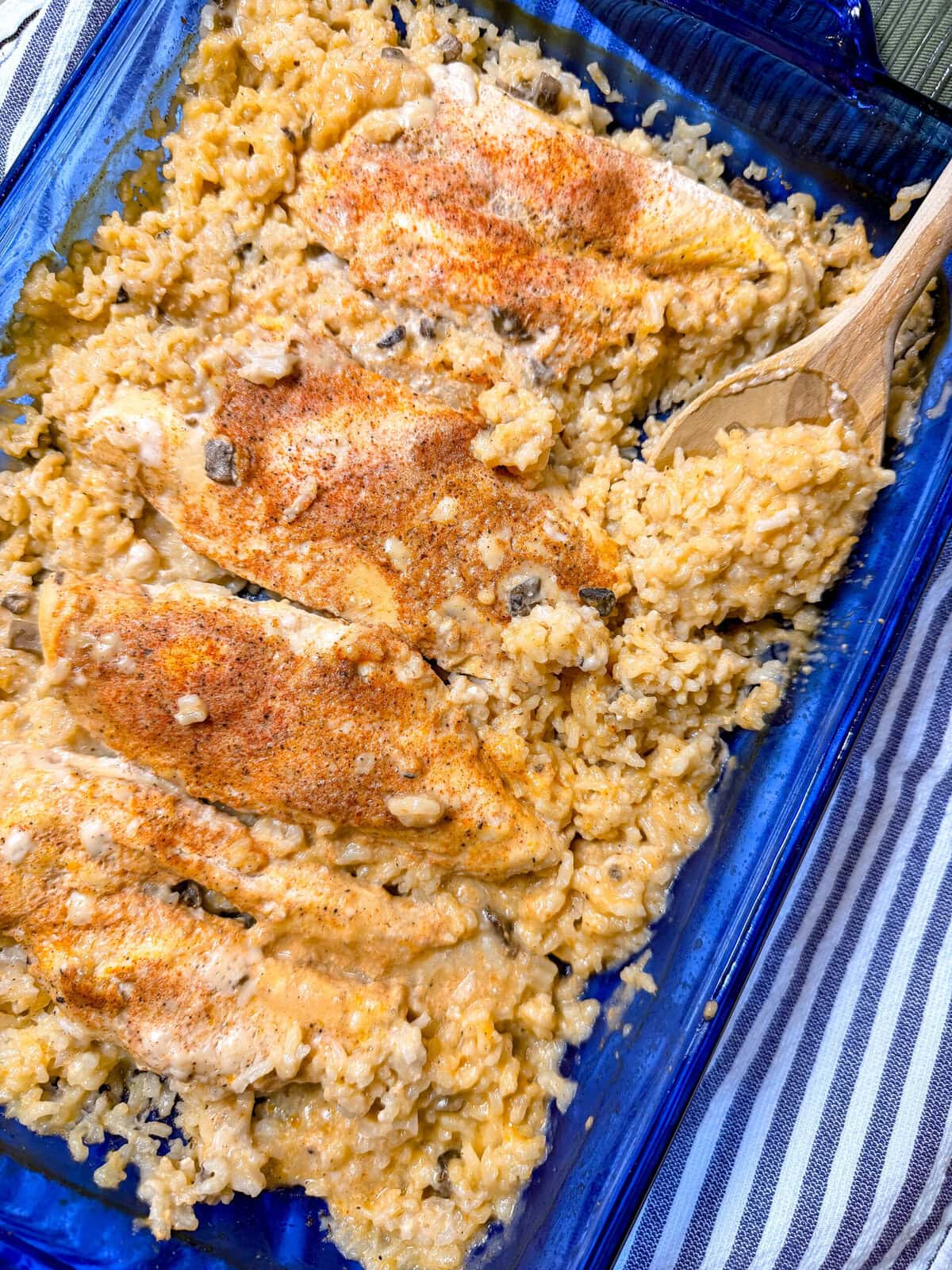 A blue baking dish filled with Creamy Chicken and Rice Casserole, topped with a sprinkle of seasoning. A wooden spoon is scooping up some of the creamy rice, and a striped cloth is partially visible underneath the dish.