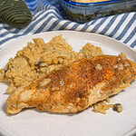 A white plate with a serving of Creamy Chicken and Rice Casserole, featuring seasoned baked chicken breast and creamy rice, with a baking dish and striped cloth in the background.