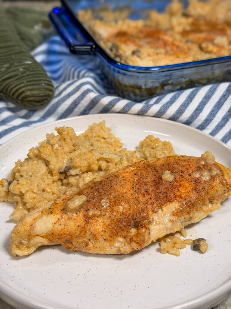A white plate with a serving of Creamy Chicken and Rice Casserole, featuring seasoned baked chicken breast and creamy rice, with a baking dish and striped cloth in the background.