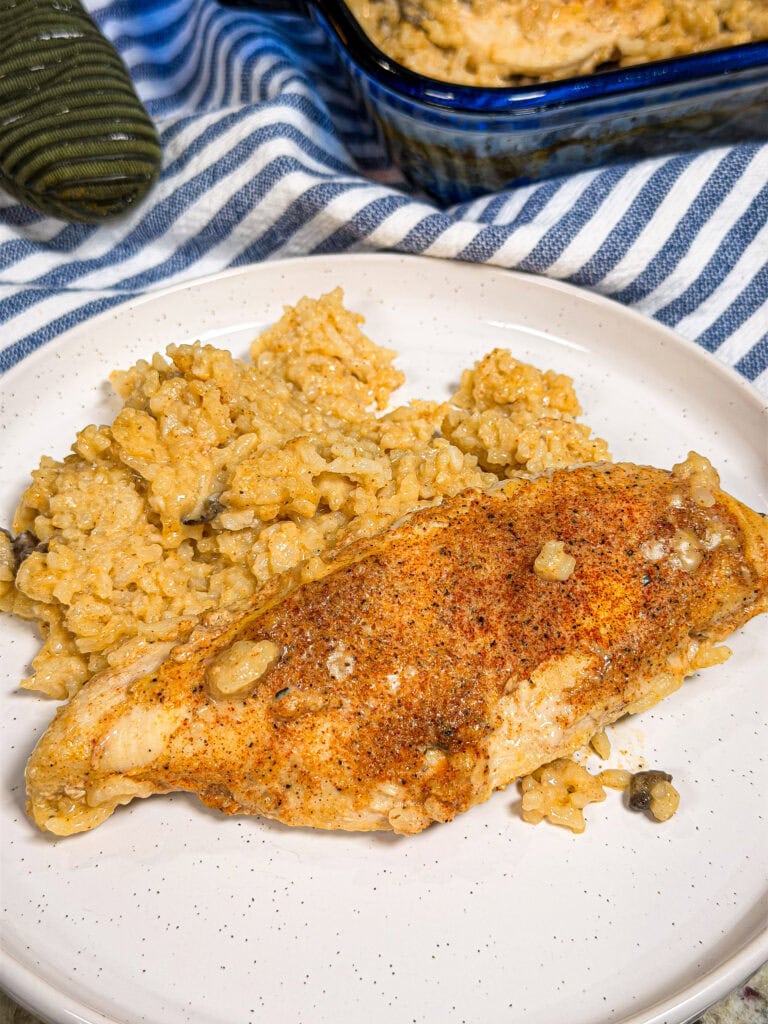 A seasoned baked chicken breast served with creamy rice on a white plate, set atop a striped cloth, evokes the comforting flavors of classic Creamy Chicken and Rice Casserole, with extra rice and chicken waiting in the background.