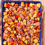 Easy Sheet Pan Pineapple Chicken features a sheet pan filled with diced chicken, pineapple, red onion, and colorful bell peppers on a blue tray, ready for baking. The tray sits on a striped kitchen towel atop a wooden surface.