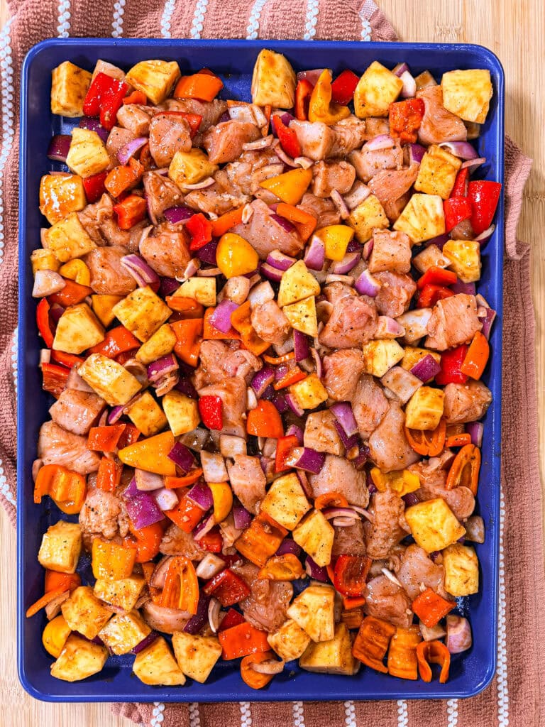 Easy Sheet Pan Pineapple Chicken features a sheet pan filled with diced chicken, pineapple, red onion, and colorful bell peppers on a blue tray, ready for baking. The tray sits on a striped kitchen towel atop a wooden surface.