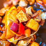 A close-up of a spoonful of cooked chicken, yellow potatoes, red onions, and red bell peppers in a savory sauce, with more of the Easy Sheet Pan Pineapple Chicken blurred in the background.