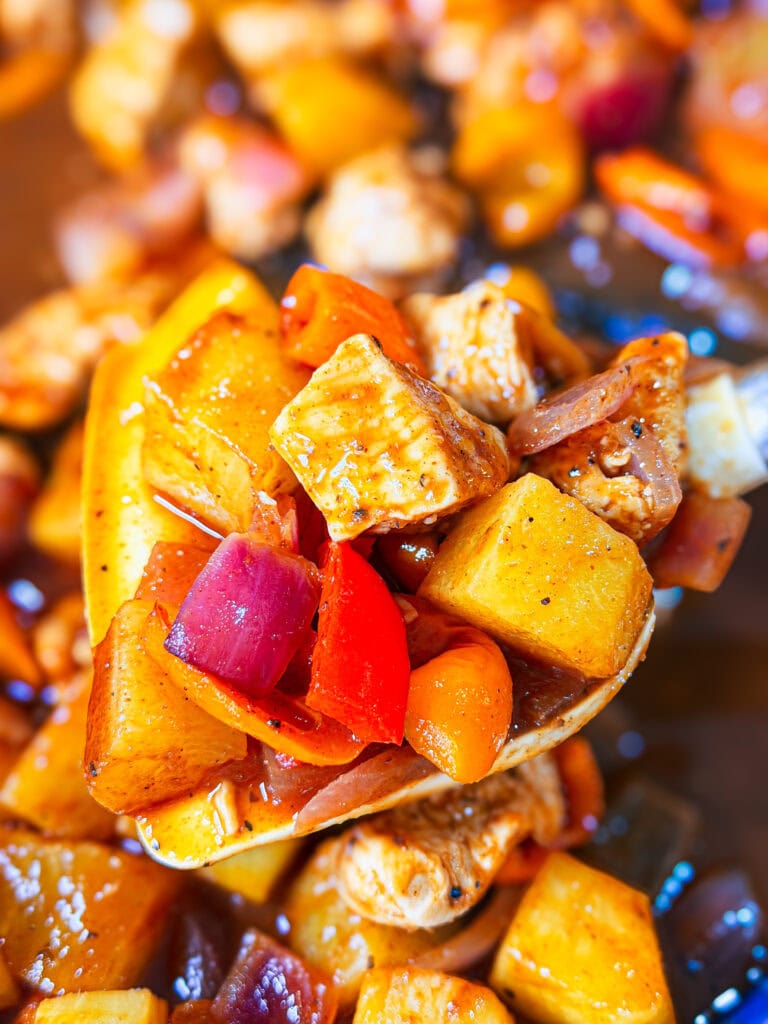 A close-up of a spoonful of cooked chicken, yellow potatoes, red onions, and red bell peppers in a savory sauce, with more of the Easy Sheet Pan Pineapple Chicken blurred in the background.