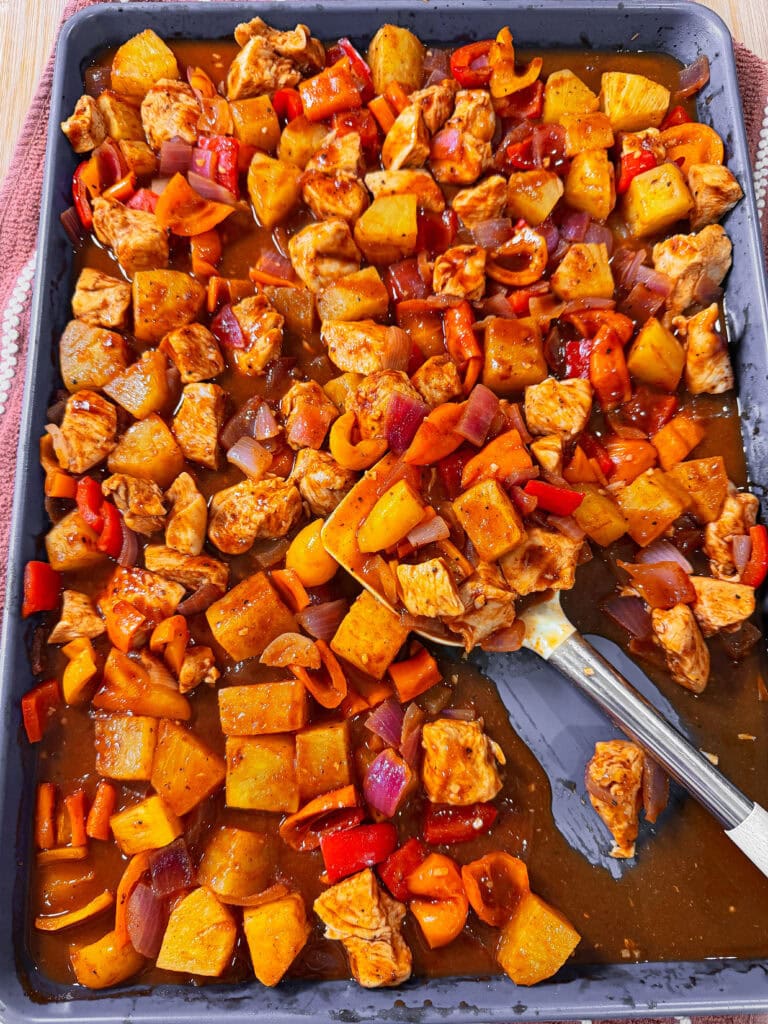 A sheet pan of Easy Sheet Pan Pineapple Chicken features baked sweet and sour chicken with chunks of pineapple, red bell peppers, onions, and tangy sauce, with a serving spoon on the right side.