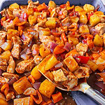 A close-up of a colorful Easy Sheet Pan Pineapple Chicken dish with chunks of chicken, potatoes, red bell peppers, carrots, and onions in a glossy brown sauce on a baking tray, with a serving spoon scooping some out.
