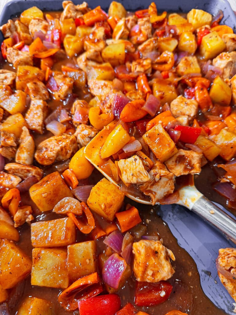 A close-up of a colorful Easy Sheet Pan Pineapple Chicken dish with chunks of chicken, potatoes, red bell peppers, carrots, and onions in a glossy brown sauce on a baking tray, with a serving spoon scooping some out.