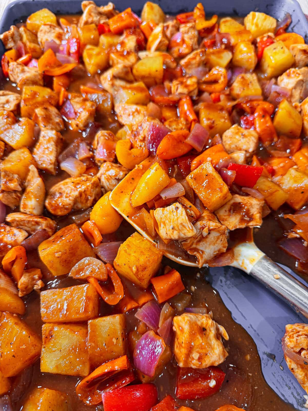 A close-up of a colorful Easy Sheet Pan Pineapple Chicken dish with chunks of chicken, potatoes, red bell peppers, carrots, and onions in a glossy brown sauce on a baking tray, with a serving spoon scooping some out.