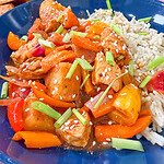 A blue plate with white rice, chunks of Easy Sheet Pan Pineapple Chicken, bell peppers, pineapple, and carrots in a savory sauce, topped with sesame seeds and chopped green onions.