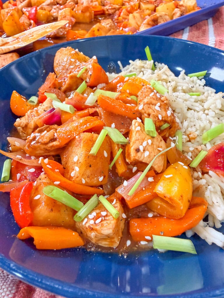 A blue plate with white rice, chunks of Easy Sheet Pan Pineapple Chicken, bell peppers, pineapple, and carrots in a savory sauce, topped with sesame seeds and chopped green onions.