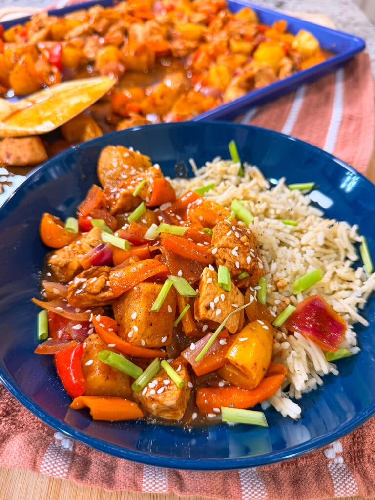 A blue plate with rice and stir-fried chicken, bell peppers, carrots, and pineapple, topped with sesame seeds and chopped green onions. In the background, a baking dish filled with more Easy Sheet Pan Pineapple Chicken adds an inviting touch to the scene.