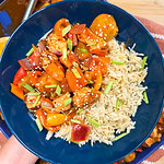 A blue bowl filled with rice, colorful stir-fried chicken and vegetables reminiscent of an Easy Sheet Pan Pineapple Chicken, topped with sesame seeds and chopped green onions; a serving spoon and more food are in the background.