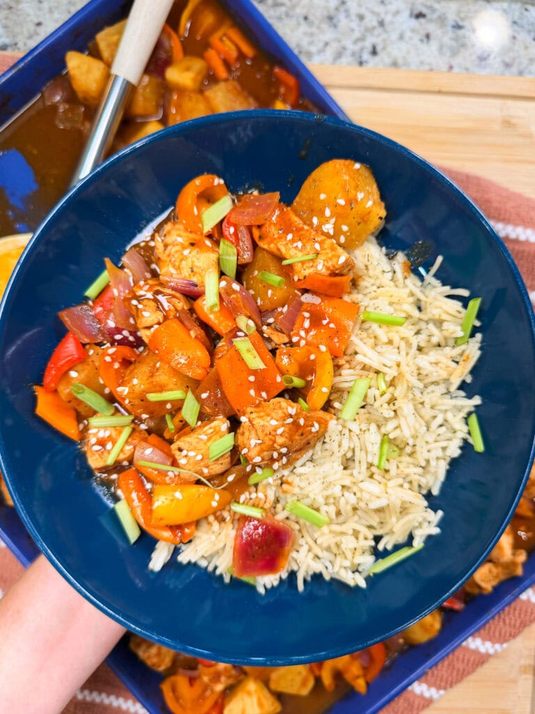 A blue bowl filled with rice, colorful stir-fried chicken and vegetables reminiscent of an Easy Sheet Pan Pineapple Chicken, topped with sesame seeds and chopped green onions; a serving spoon and more food are in the background.