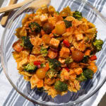 A glass bowl filled with The Best Pasta Salad—bowtie pasta, cherry tomatoes, broccoli, red peppers, tofu cubes, and sprinkled spices—sits on a blue and white striped cloth.