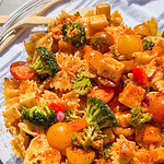 A close-up of The Best Pasta Salad featuring bowtie pasta, broccoli florets, cherry tomatoes, cheese cubes, and red bell peppers, all seasoned with herbs and spices in a glass bowl.